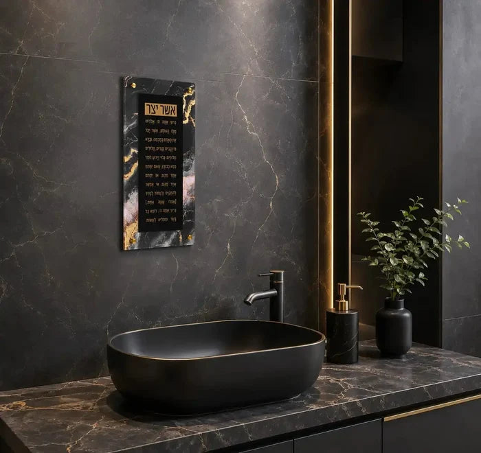Black agate floating wall art displaying Asher Yatzar text, mounted above a modern bathroom sink with gold accents and marble countertop.