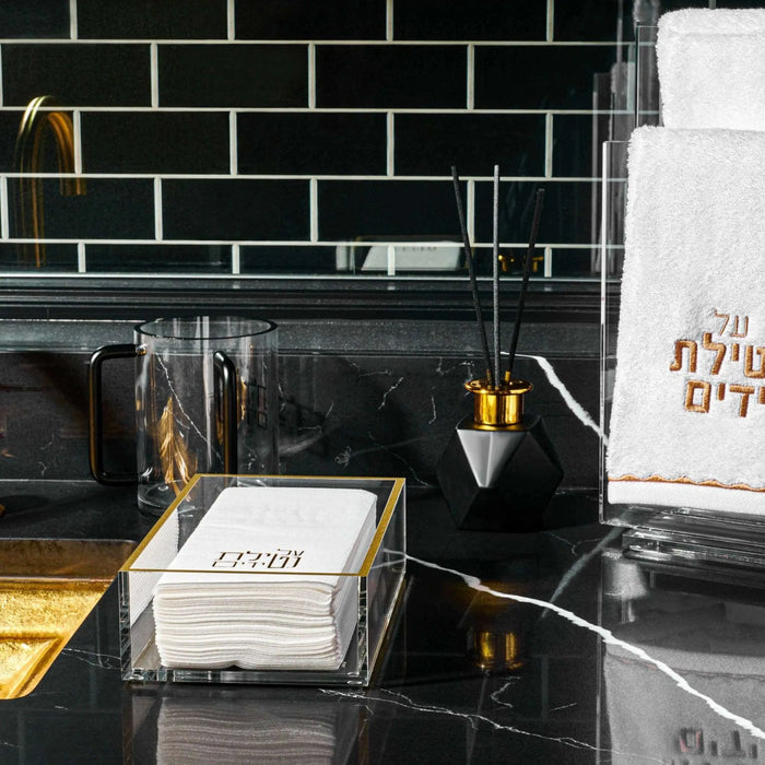 Elegant white ceramic Netilas Yadayim washing cup displayed on a black bathroom countertop with gold accents and Jewish prayer books nearby.