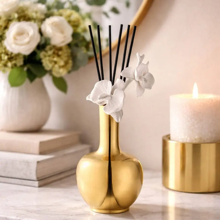 Gold pear-shaped Aura Orchid Vase Diffuser with black reeds and white orchid flowers, styled with candles and books on a white surface.
