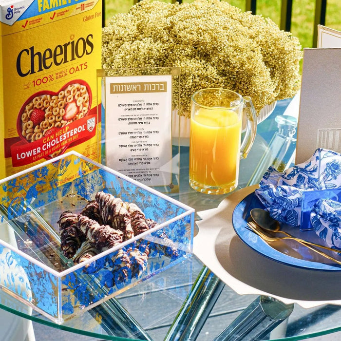 Basics Brachos Rishonos tabletop display with blue tile holder, cereal box, and juice glass on breakfast table setting.