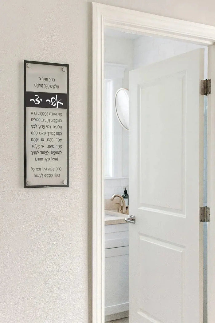 Black and gold Asher Yatzar prayer wall art hanging beside a white bathroom door, displaying traditional Jewish blessing text in an elegant frame.
