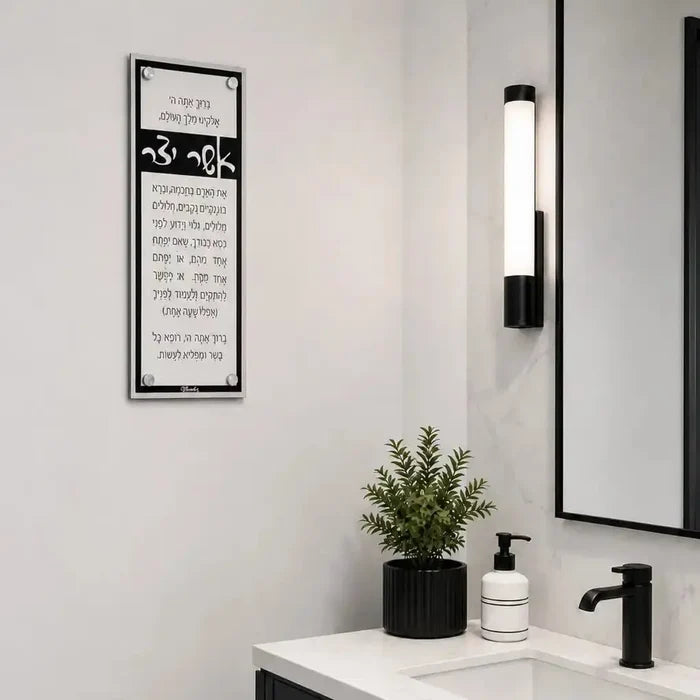 Black framed Asher Yatzar prayer wall art hanging above a modern bathroom sink with black fixtures and minimalist decor