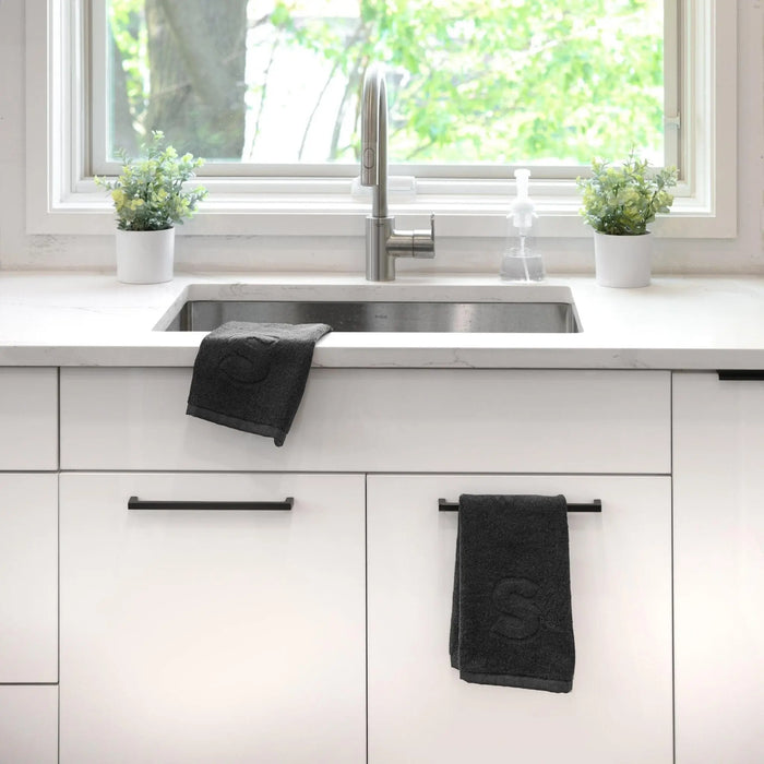 Black finger towel with embossed initial monogram displayed on bathroom vanity sink counter next to white cabinetry and window.