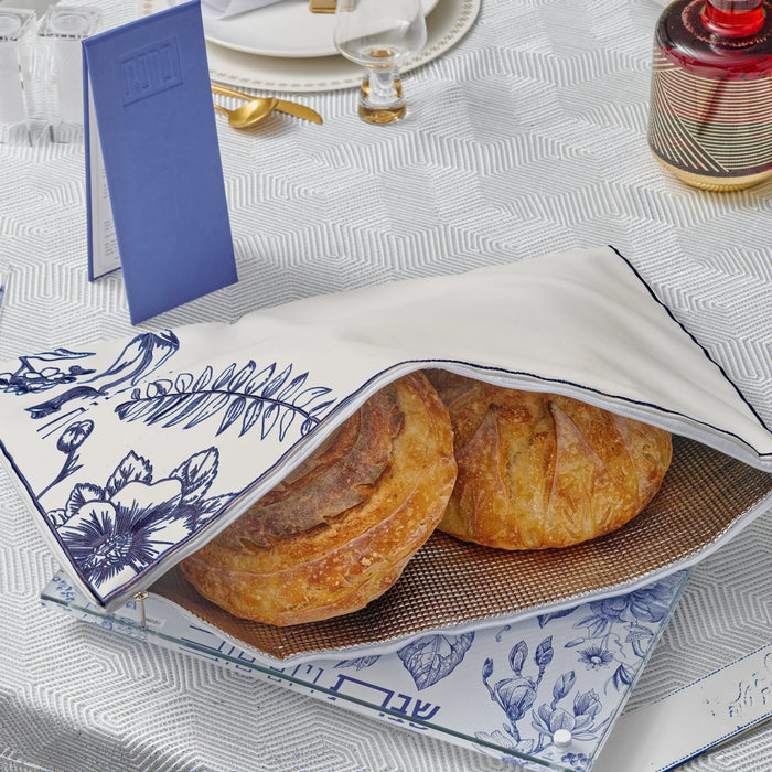 French Toile Challah Pouch Cover in white and gold holding two braided loaves on a table setting with blue toile napkin
