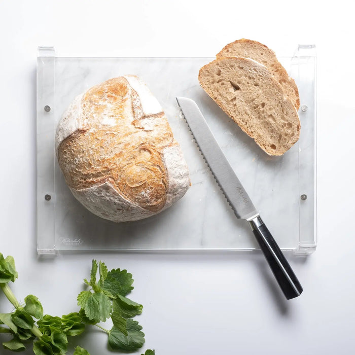 Waterdale Collection bread knife with serrated blade next to sliced crusty bread on white cutting board with fresh parsley.