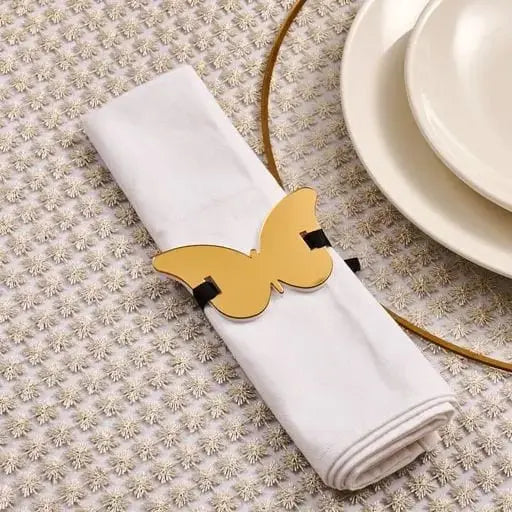 Gold butterfly napkin wrap holding a white linen napkin on a beige textured tablecloth next to white plates.