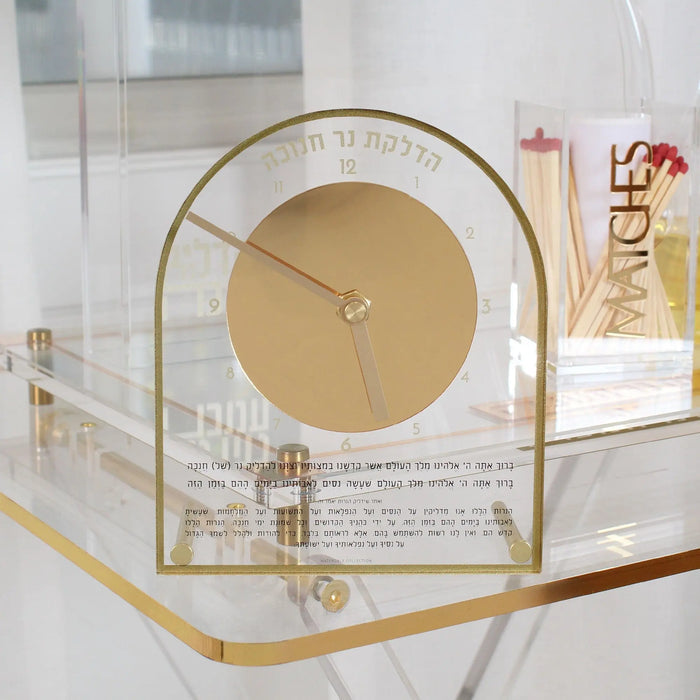 The Chanukah Brachos Clock in gold, set out with Chanukah pieces on a white surface, its brachos printed beneath the dial