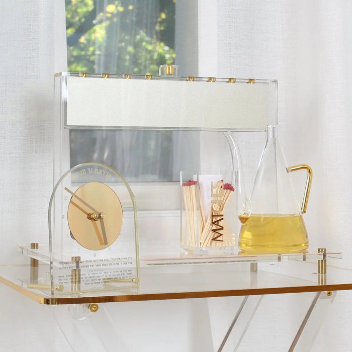 Suspended Menorah with clear acrylic frame and brass accents holding candles, a Chanukah Brachos tabletop clock, and a glass oil jug, displayed on white shelf near window with natural lighting.
