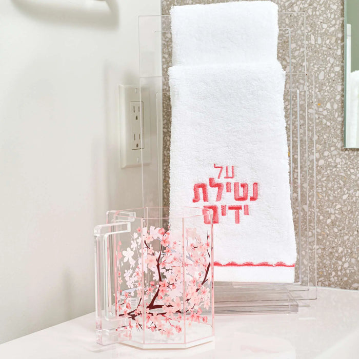 White scalloped finger towel with pink Hebrew text, displayed next to a clear glass on a bathroom vanity.