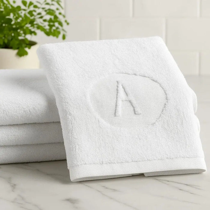 White embossed finger towel with circular initial monogram design, folded and stacked with additional towels, perfect for bathroom vanity or gifting.