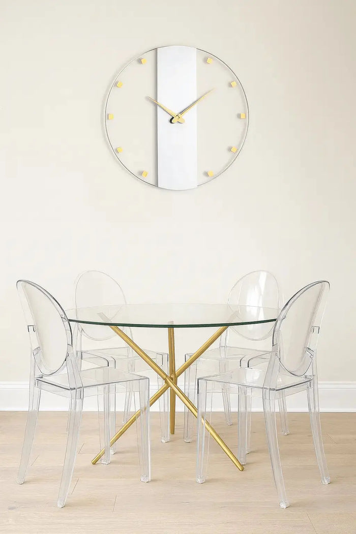 Classic minimalist wall clock with brass accents and clear face displayed above a modern dining setup with transparent chairs.