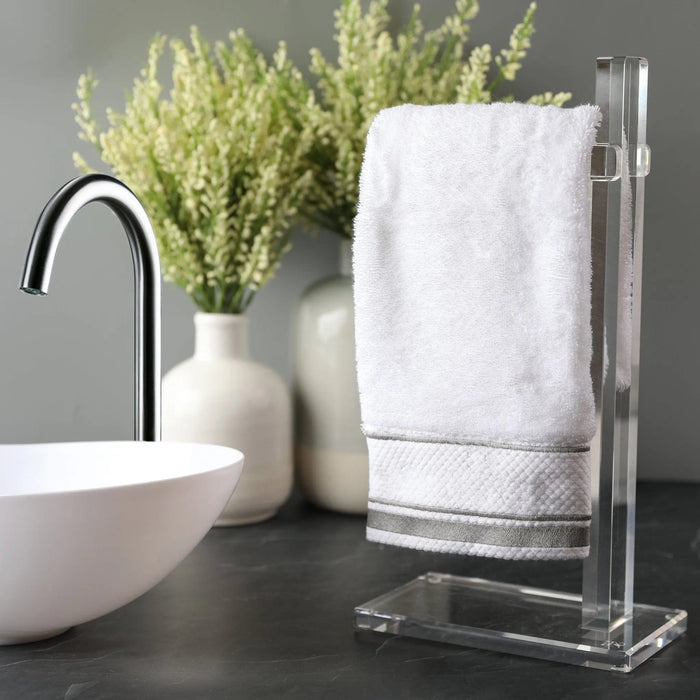 Classic clear acrylic finger towel stand holding a white towel next to a modern white sink basin and decorative plant arrangement on bathroom vanity.