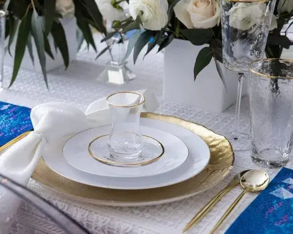 Classic glass cups and saucers with gold trim displayed on formal tabletop setting with blue runner and floral arrangement.