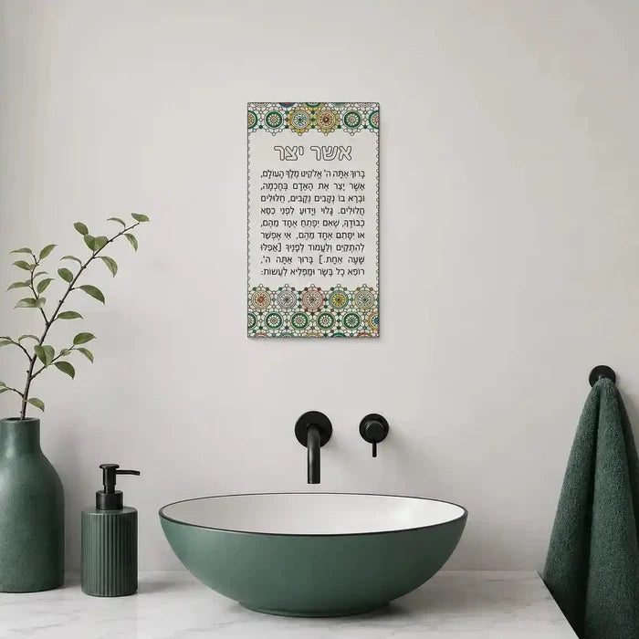 Green lucite Asher Yatzar prayer card with ornate border displayed above marble bathroom sink with black fixtures and green accessories.