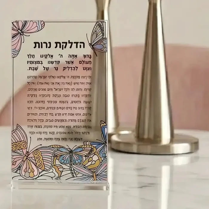 Clear acrylic Hadlokas Neiros card with butterfly design and Hebrew text, displayed next to silver candlestick holders for Shabbat candle lighting.