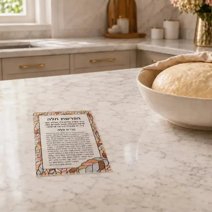 Colorful acrylic hafrashas challah card with Hebrew text and decorative bread design, displayed on marble kitchen counter next to fresh challah bowl.