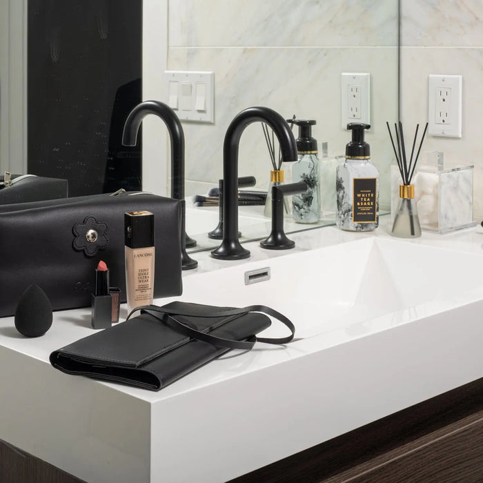 Black leather cosmetic bag by Waterdale Collection displayed on bathroom counter next to skincare products and black faucet