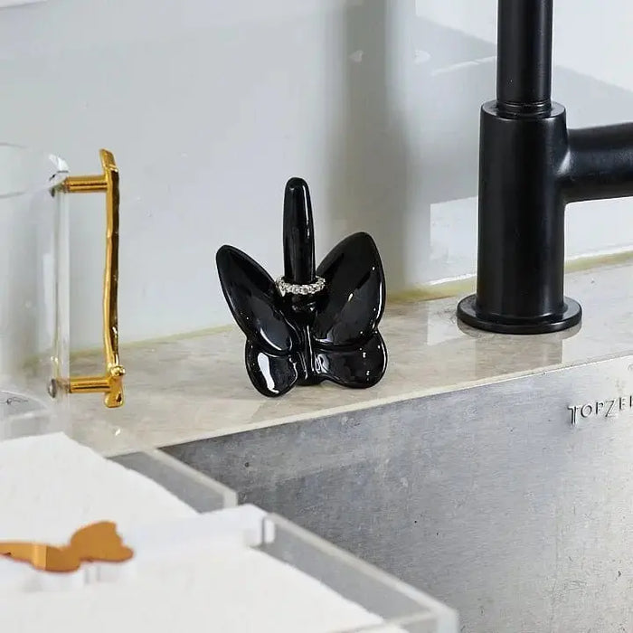 Black crystal butterfly ring holder displayed on bathroom counter with gold and black faucet accessories nearby