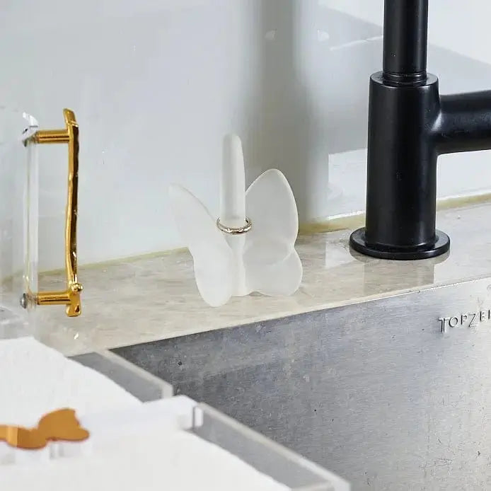Crystal butterfly ring holder in white displayed on a marble bathroom counter with gold and black faucet accessories nearby.