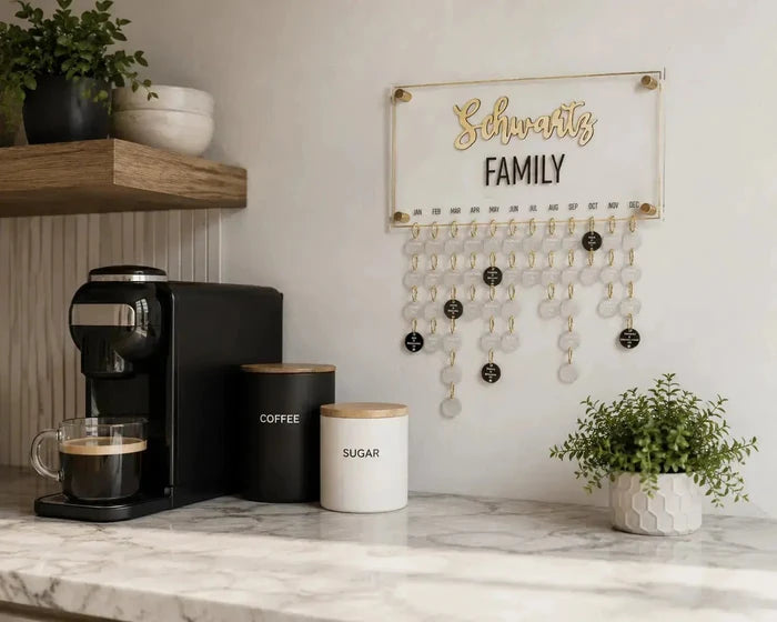 Custom family occasion calendar with hanging charms displayed on white wall above kitchen counter with coffee maker and plants.