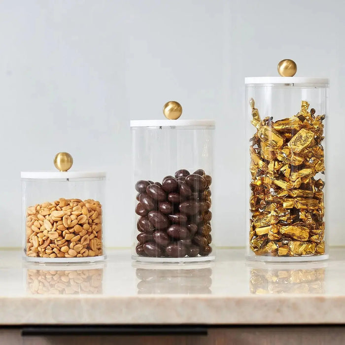 Three clear cylinder canisters in graduating sizes with brass lids, filled with cereal, chocolate candies, and popcorn on a marble surface.
