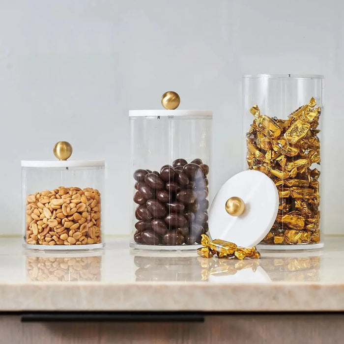 Four clear cylinder canisters with brass lids holding cereal, chocolate chips, pasta, and cornflakes on a marble surface.