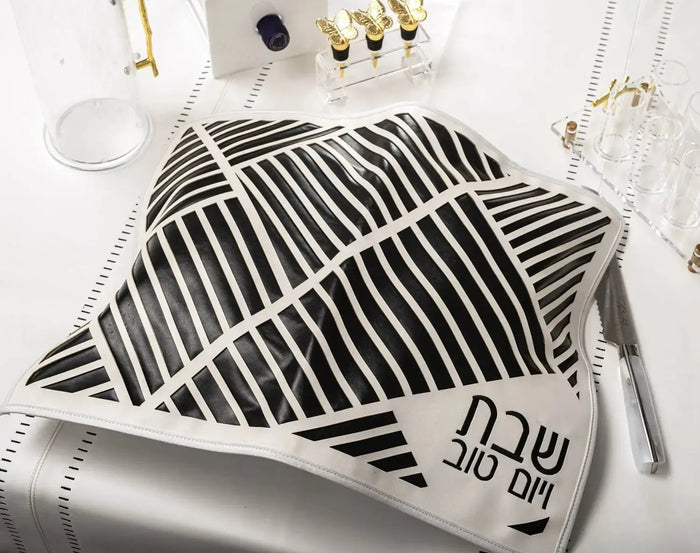 Black leather challah cover with diagonal laser-cut geometric pattern, displayed with Shabbat tabletop items and gold accents