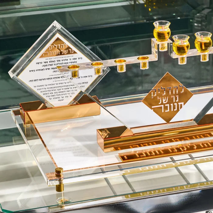 The Diamond Chanukah Brachos Tabletop in its gold base beside a clear Lucite menorah, set out on glass for Hanukkah lighting