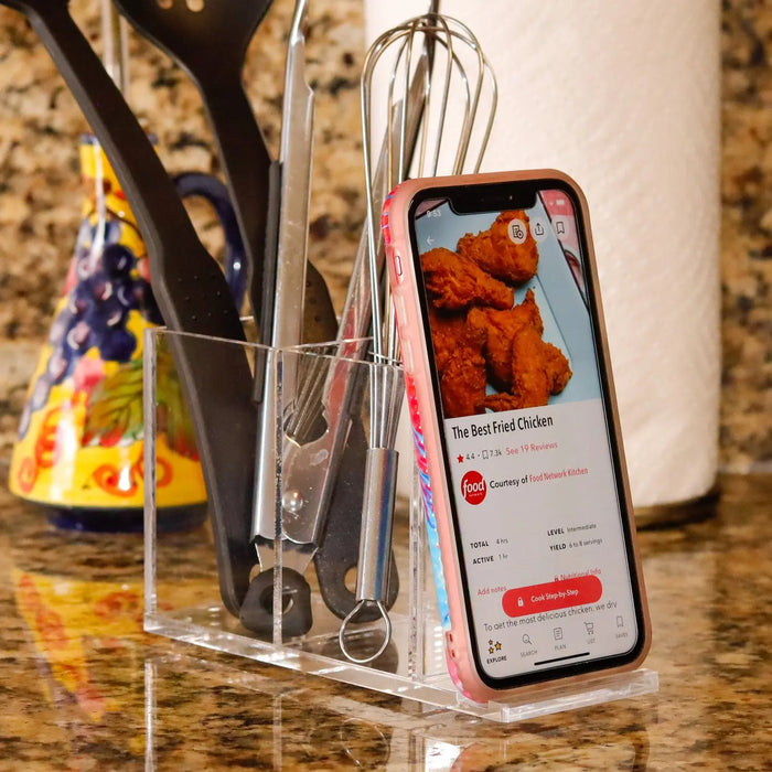 Black metal double pen and phone holder displaying smartphone with recipe app and storing pens on marble surface near plant.