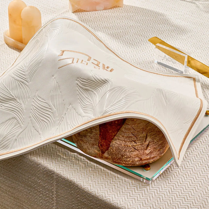 Embossed leaf design on gold and silver challah board with leather accents, displayed on white linens with shabbat items