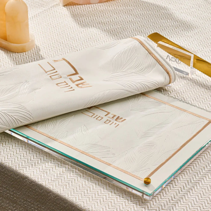 Embossed leaf challah board in gold and black leather by Waterdale Collection, displayed with a white napkin and gold utensils on a textured surface.