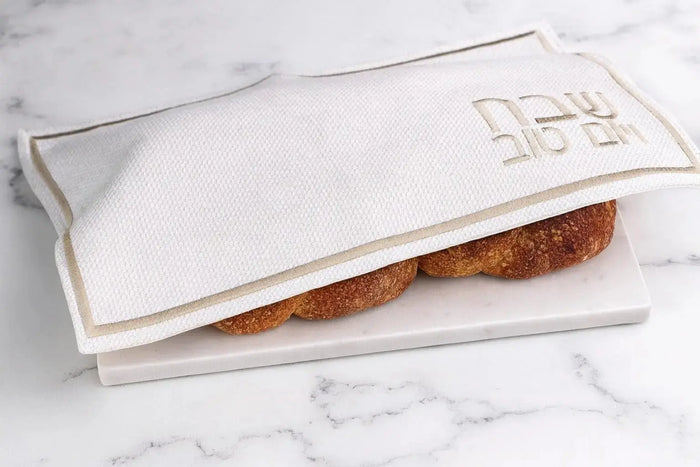 Cream and gold fabric challah cover with embroidered design, displayed open on marble surface with challah bread visible inside