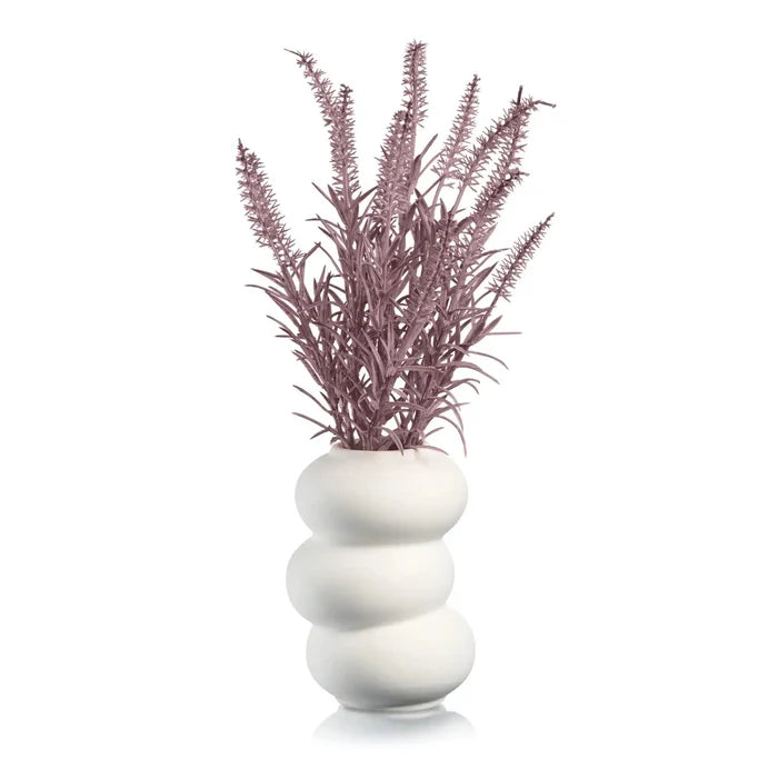 White ceramic donut vase with stacked ring design holding dried purple faux flowers and grasses arrangement.