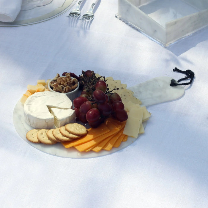 Flat marble charcuterie board topped with cheese, crackers, and grapes, displayed on white surface with serving utensils nearby.