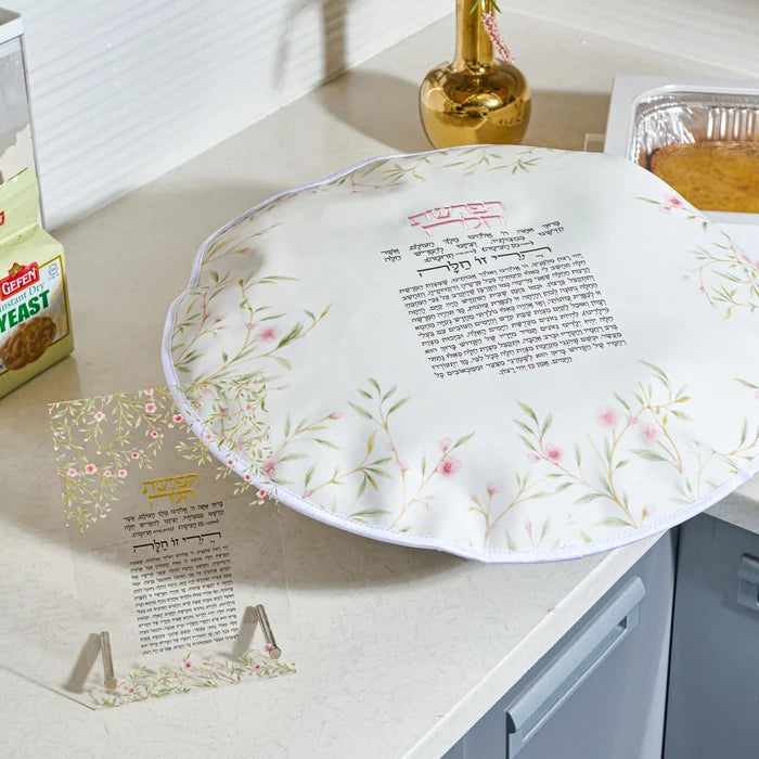 A round white floral challah dough cover with a floral border and printed Hebrew text, displayed on a kitchen countertop next to a  clear acrylic cup and a box of yeast.