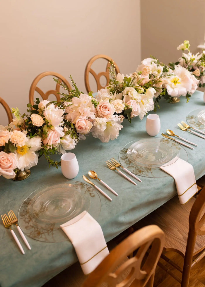 Silver floral chargers with peach roses, greenery, and woven handles arranged on a sage green table runner with place settings.