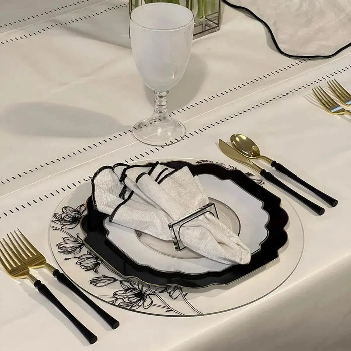 Black floral charger plate with silver border on white tablecloth, set with white plate, black cutlery, and wine glass for formal dining.
