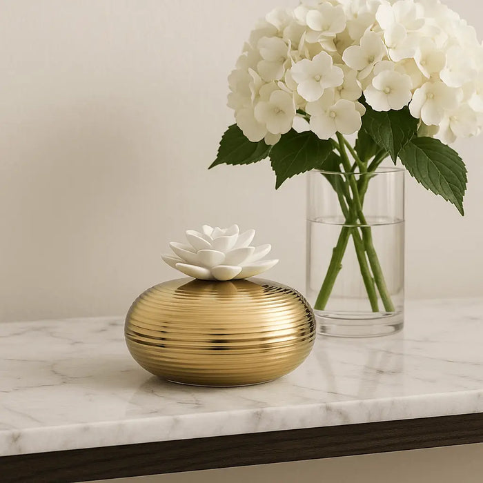 Gold fluted scent diffuser beside white hydrangeas in a vase, styled on a marble surface.