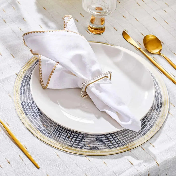 Geometric charger plate with gray and gold circular pattern, white napkin with beaded trim, and gold utensils on white textured surface.