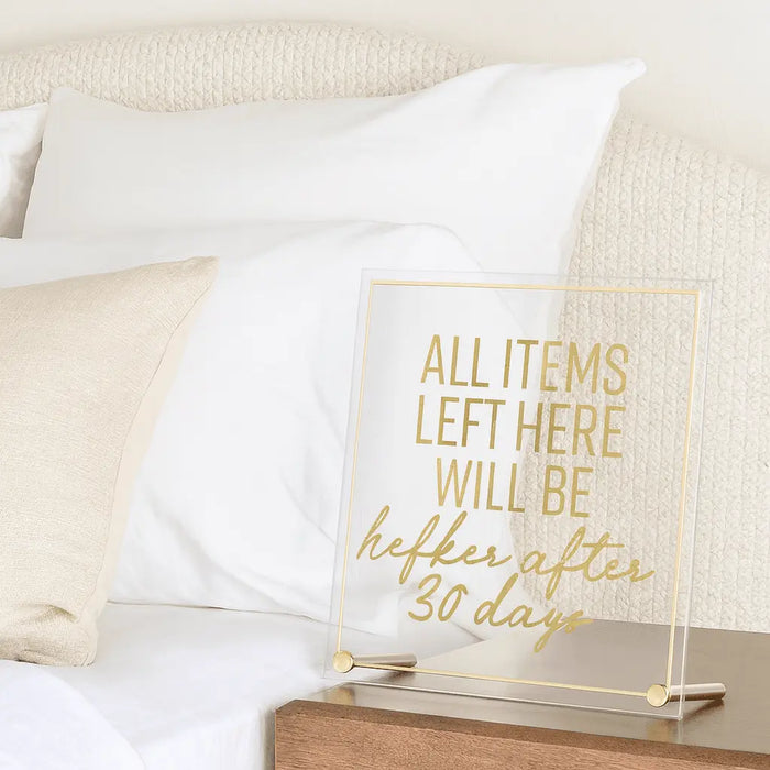 Wooden guest room sign with gold foil text reading "All items left here will be donated after 30 days" displayed on nightstand next to bed.
