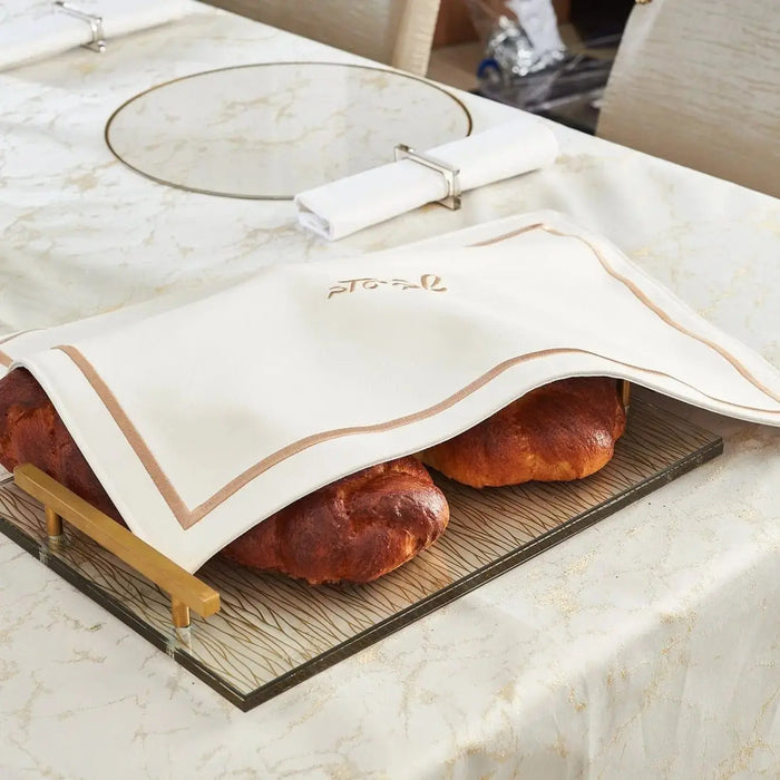 White leather challah cover with gold embroidery displayed on marble surface with fresh challah bread underneath, from Waterdale Collection.
