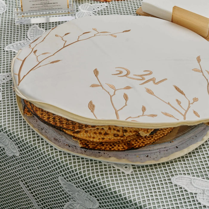 White and gold leaf embroidered matzah pouch cover with leather trim, displayed on patterned fabric for Pesach.