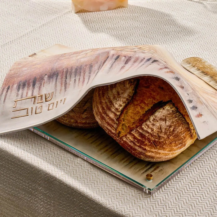 Taupe leather Jerusalem Challah Cover with embossed Kosel design, displayed open over fresh baked challah bread on white surface.