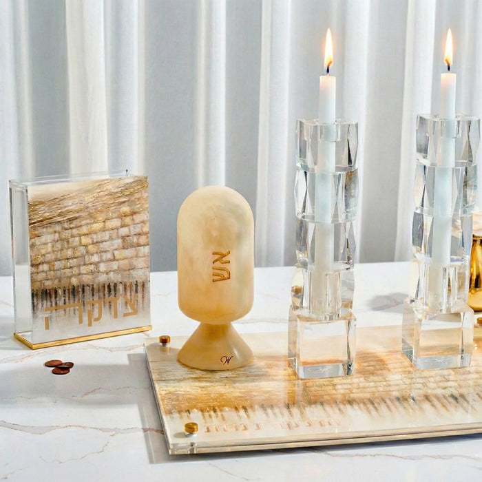 Taupe Jerusalem Tzedakah box with painted Western Wall design, displayed with candles and gold accessories on marble surface.