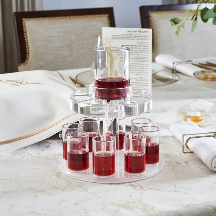 Clear glass Kiddush fountain with multiple tiers holding small wine cups, displayed on a marble table with Shabbat dinnerware and place settings.