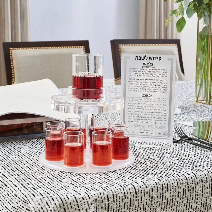 Waterdale Collection Kiddush fountain with red wine in glass cups arranged on a grey tablecloth, displayed on a bedroom nightstand with Shabbat prayer book.