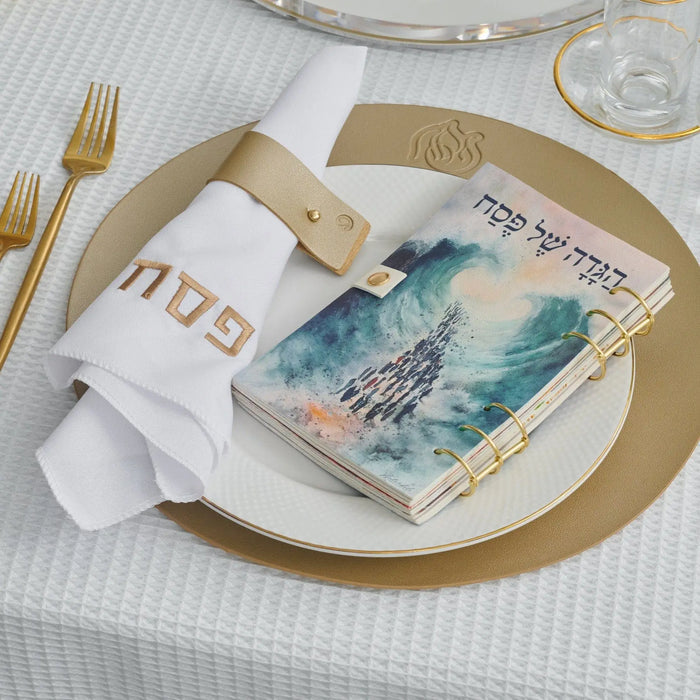 Kids leather painted Haggadah in blue displayed on white plate with gold utensils and napkin at Pesach table setting.