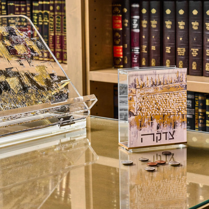 Kosel at Winter Tzedakah Box displayed on marble surface with bookshelves in background, featuring painted winter landscape design