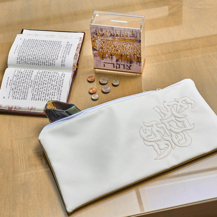 White Kosel at Winter tzedakah box with embroidered Hebrew text, displayed with prayer book, coins, and religious items on neutral surface.
