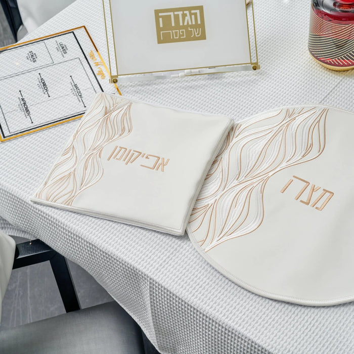 White leather Lace Afikoman Bag with gold embroidered details, displayed on tabletop with Passover items and design sketches nearby.